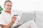 Happy man on his couch using laptop for shopping online looking at camera