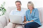 Happy couple using laptop together on the couch looking at camera at home in living room