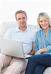 Couple using laptop together on the couch smiling at camera at home in living room