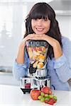 Smiling brunette leaning on her juicer full of fruit looking at camera in kitchen