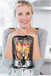Blonde woman leaning on her juicer full of fruit and smiling at camera at home in kitchen