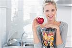 Happy woman leaning on her juicer full of fruit and holding red apple looking at camera at home in kitchen