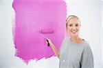 Smiling blonde painting her wall pink in her new home looking at camera