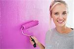 Cheerful blonde painting her wall pink and smiling at camera in new home