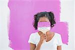 Happy woman hiding her face with paintbrush againstly freshly painted pink wall
