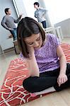 Girl sitting on floor with parents arguing in background