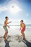 Men in swim trunks heading soccer ball on beach