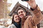 Smiling couple taking self-portrait with digital camera outside snowy cabin