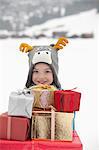 Portrait of smiling boy wearing reindeer hat and carrying stack of Christmas gifts in snow