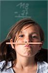 Portrait of girl standing in front of blackboard in classroom, holding pencil with mouth, Germany