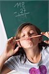 Portrait of girl standing in front of blackboard in classroom, holding pencil over mouth, Germany