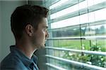 Close-up portrait of young man, looking out window through blinds, Germany