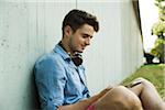 Young man sitting next to wall of building outdoors, with headphones around neck and looking at cell phone, Germany