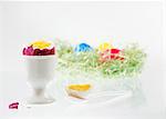 A boiled egg with the top cut off, in an eggcup, with an Easter nest of colourful eggs in the background