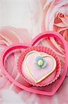 close up of one pink iced, love heart shaped cup cake with a pink plastic heart shape biscuit cutter