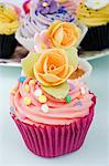 still life of a row of coloured cup cakes decorated with orange rose flowers on top in their cake papers on a white table with other cup cakes in the background
