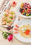 A breakfast of a caviar-topped waffle, slices of bread topped with cheese and ham, fresh fruits and a strawberry and mango smoothie