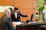 Receptionist pointing on a desktop pc with a businessman at the hotel reception counter