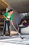 Woman cleaning house with a vacuum cleaner with her husband sitting on a seat