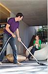 Man cleaning house with a vacuum cleaner with his wife sitting on a seat