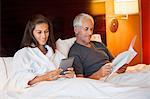 Woman using a digital tablet with her husband reading a book in a hotel room