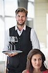 Woman sitting in a restaurant with a waiter holding a tray of wine glasses in the background