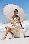 Beautiful woman sitting on a suitcase with an umbrella on the beach