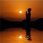 Silhouette woman sitting and relaxing against orange sunset with reflection in water