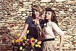 couple of fashion women with vintage style posing near basket of the bicycle with some colourful flowers near a stone wall , in vintage color