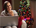 Smiling young woman with laptop near christmas tree looking on copy space