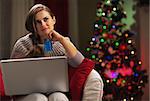 Thoughtful young woman with laptop and credit card near christmas tree