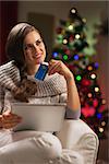 Thoughtful young woman with credit card and tablet pc near christmas tree