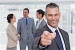 Cheerful businesman in bright office pointing at camera with colleagues on background