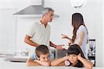 Unhappy siblings sitting in kitchen with their parents who are fighting at home loudly