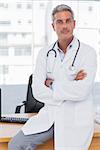 Serious doctor sitting on desk with arms crossed and looking at camera