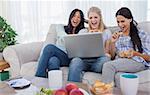 Smiling friends looking at laptop together and eating cookies at home on couch