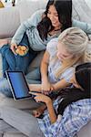 Smiling friends using digital tablet together and eating cookies at home on the couch