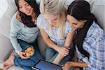 Laughing friends using digital tablet together and eating cookies at home on couch