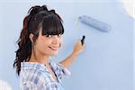 Pretty woman painting wall blue and smiling at camera in new home