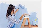 Woman using paint roller to paint wall in blue paint leaning on ladder