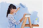 Woman using paint roller to paint wall and smiling at camera leaning on a ladder