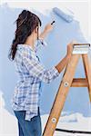 Woman using paint roller to paint wall blue standing on ladder