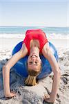Fit blonde stretching back on exercise ball smiling at camera on the beach