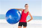 Fit blonde holding exercise ball smiling at camera on the beach