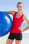 Fit woman holding exercise ball smiling at camera on the beach