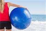 Fit blonde holding exercise ball looking at waves on the beach