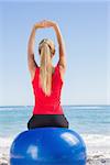 Fit blonde sitting on exercise ball looking at sea stretching arms on the beach
