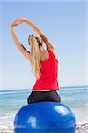 Fit woman sitting on exercise ball looking at sea stretching arms the beach