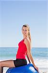 Cheerful woman sitting on exercise ball looking at camera on the beach