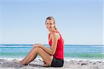 Fit blonde sitting on sand smiling at camera on the beach